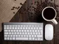 Tastiera PC con tasti funzione, mouse e tazzina di caffè su un tavolo di legno rustico, circondata da chicchi di caffè.