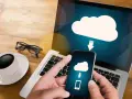 Mani che tengono uno smartphone con un'icona cloud e freccia, mostrando come salvare dati sul computer. Tazza di caffè e occhiali sul tavolo.