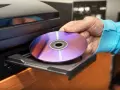 Mano inserisce un CD-ROM nel lettore. Il CD-ROM, con riflessi viola e blu, è pronto per essere letto, mostrando il suo significato di supporto di memoria.