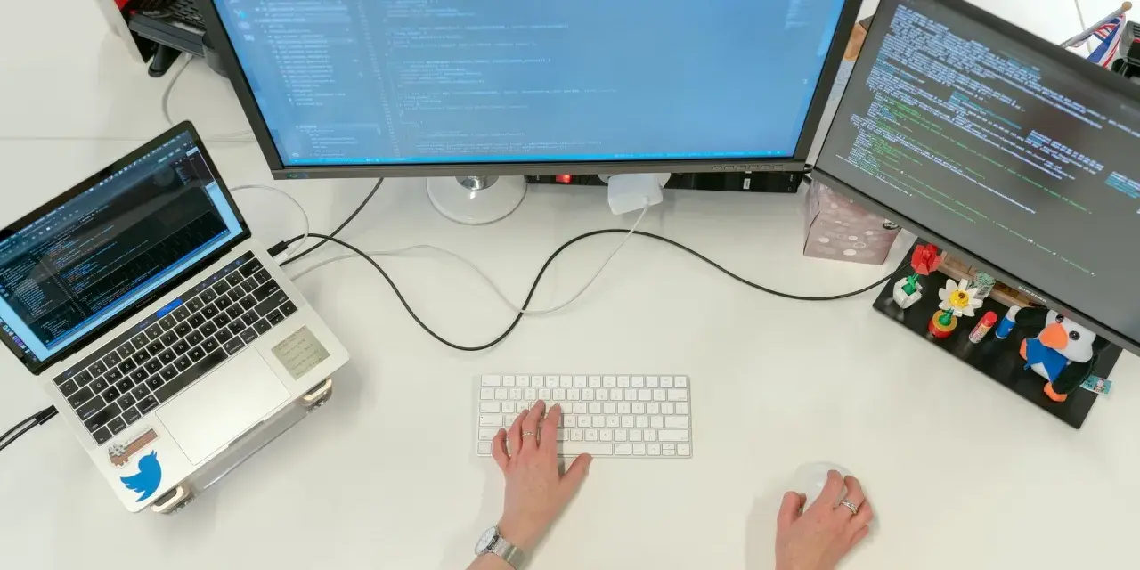 Postazione di lavoro con laptop e due monitor che mostrano codice. Una persona digita, concentrata sul suo lavoro con python, per continuare a sviluppare.