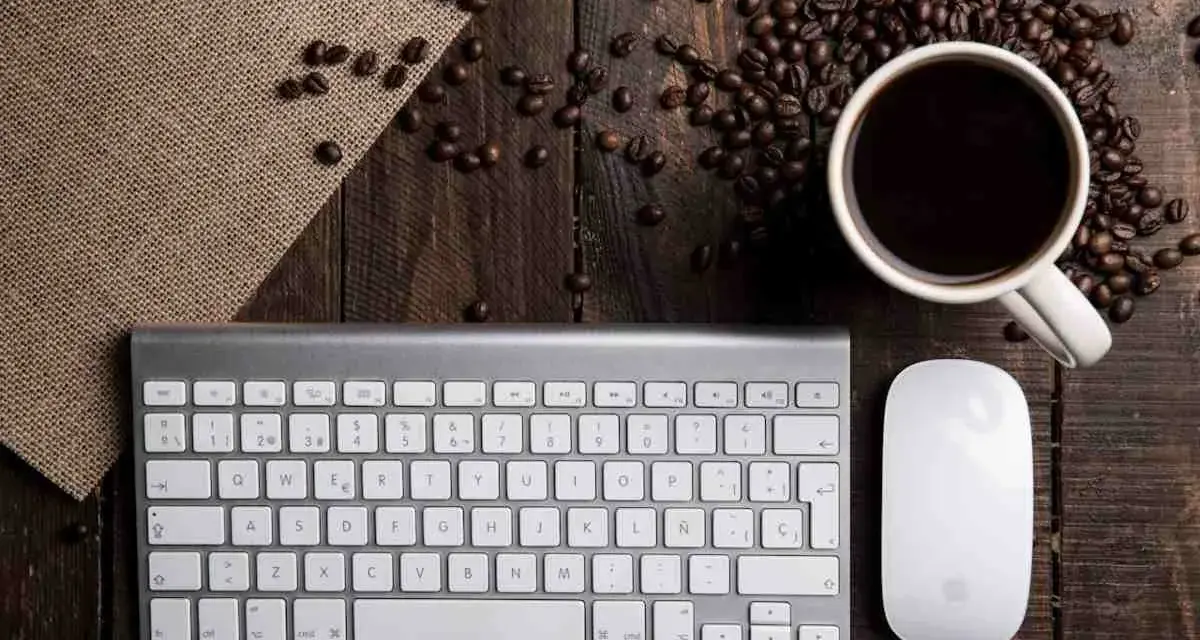Tastiera PC con tasti funzione, mouse e tazzina di caffè su un tavolo di legno rustico, circondata da chicchi di caffè.