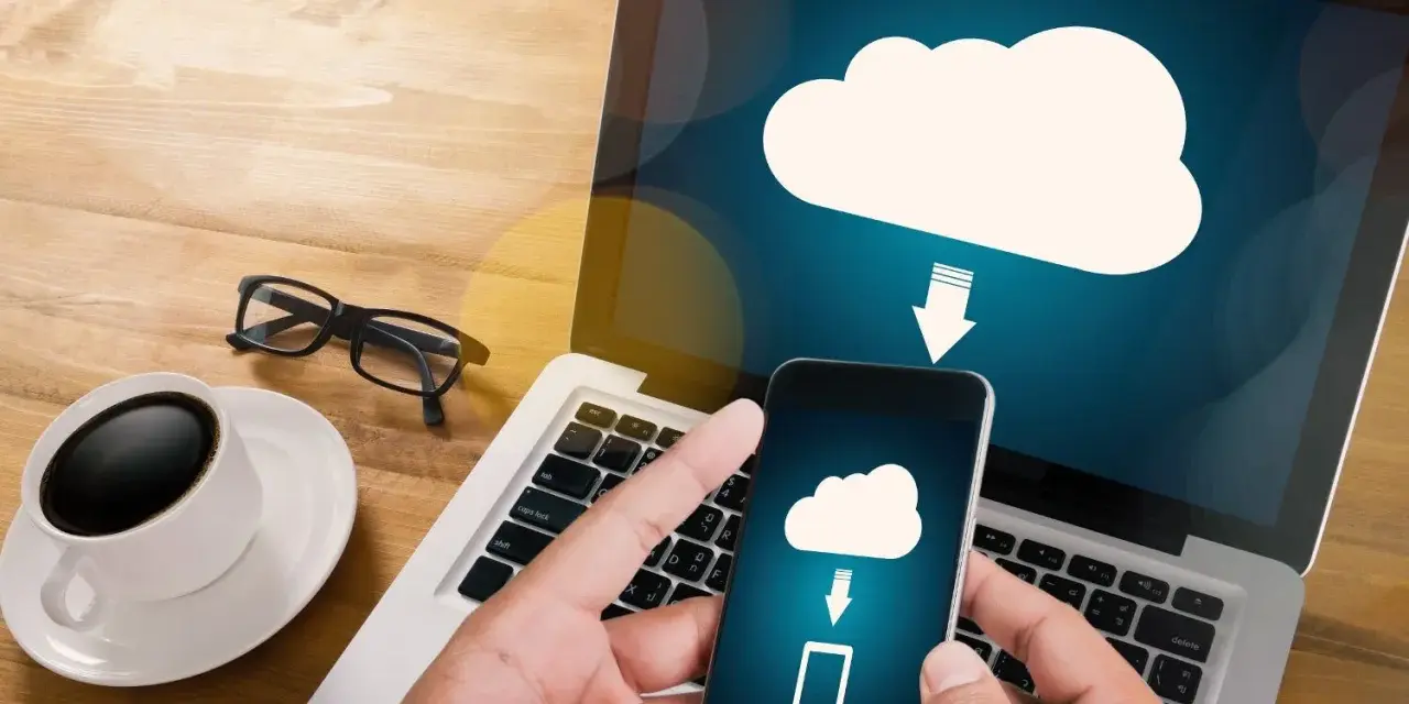 Mani che tengono uno smartphone con un'icona cloud e freccia, mostrando come salvare dati sul computer. Tazza di caffè e occhiali sul tavolo.