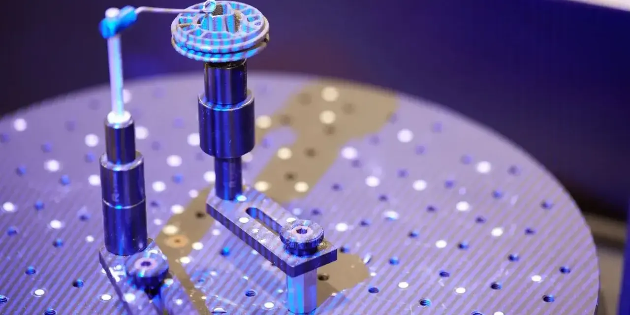 Un oggetto circolare con scanalature è fissato su un supporto. Un braccio metallico con una punta blu si avvicina per analizzarlo. Questo è un esempio di come funziona uno scanner 3D.