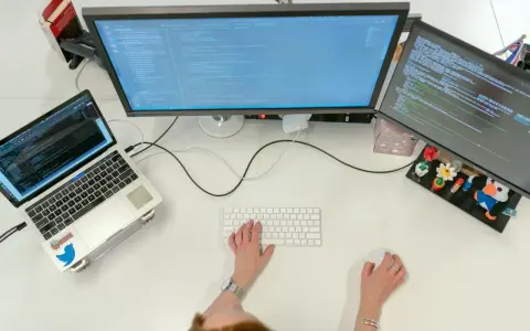 Postazione di lavoro con laptop e due monitor che mostrano codice. Una persona digita, concentrata sul suo lavoro con python, per continuare a sviluppare.
