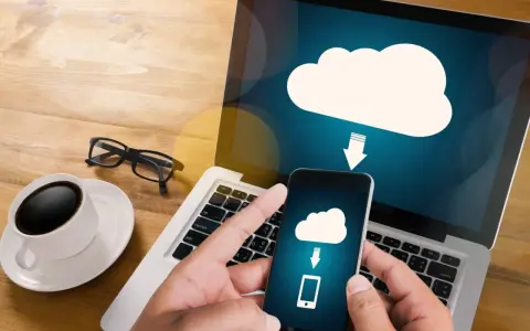 Mani che tengono uno smartphone con un'icona cloud e freccia, mostrando come salvare dati sul computer. Tazza di caffè e occhiali sul tavolo.