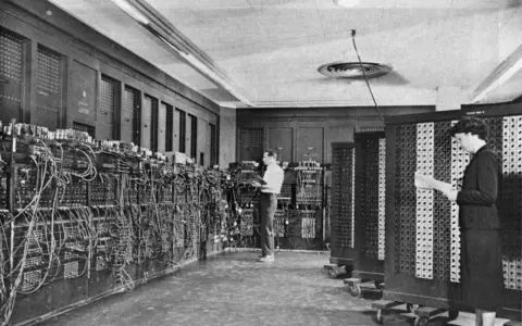 Il computer ENIAC, un gigante di cavi e pannelli, con due persone che lavorano al suo interno.