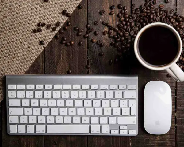Tastiera PC con tasti funzione, mouse e tazzina di caffè su un tavolo di legno rustico, circondata da chicchi di caffè.
