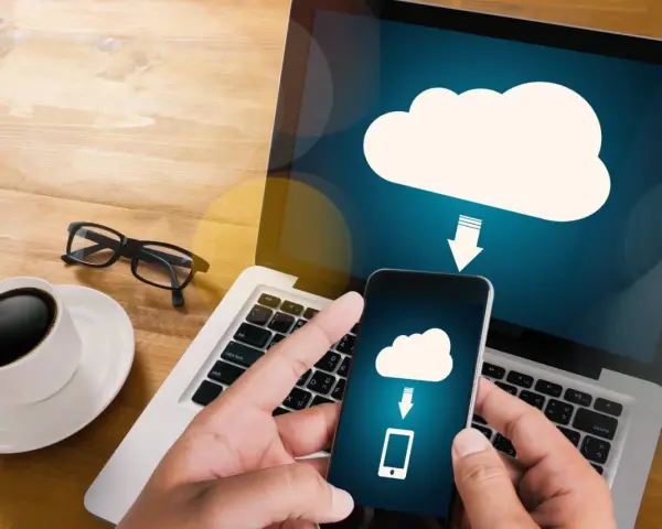 Mani che tengono uno smartphone con un'icona cloud e freccia, mostrando come salvare dati sul computer. Tazza di caffè e occhiali sul tavolo.