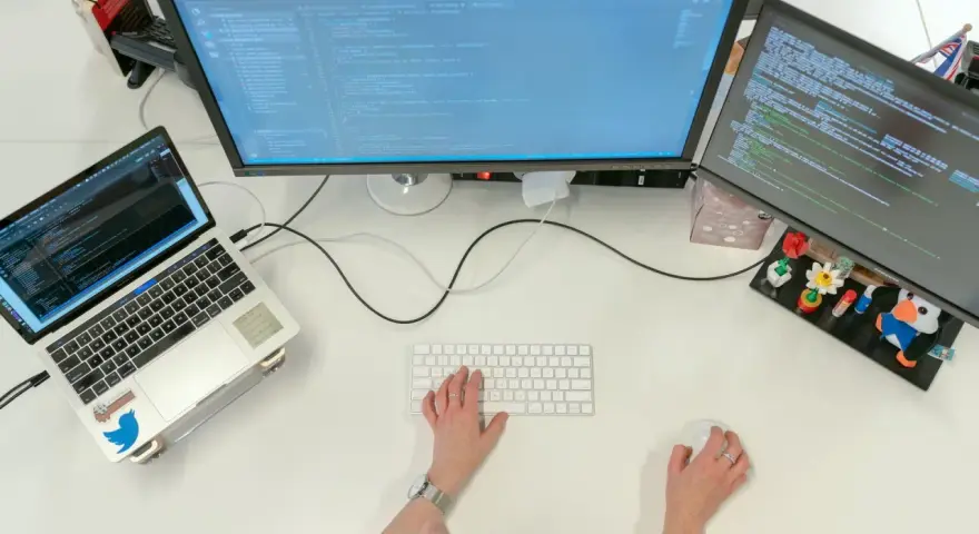 Postazione di lavoro con laptop e due monitor che mostrano codice. Una persona digita, concentrata sul suo lavoro con python, per continuare a sviluppare.