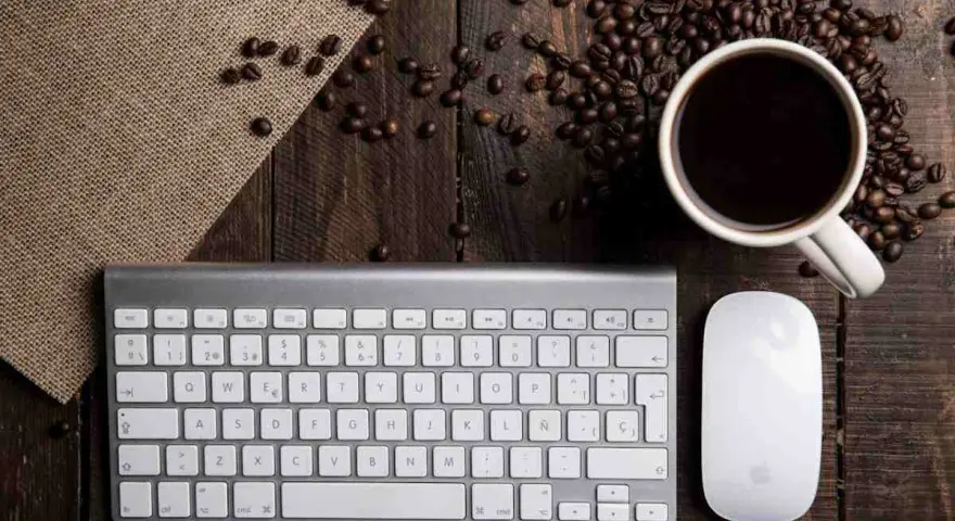 Tastiera PC con tasti funzione, mouse e tazzina di caffè su un tavolo di legno rustico, circondata da chicchi di caffè.