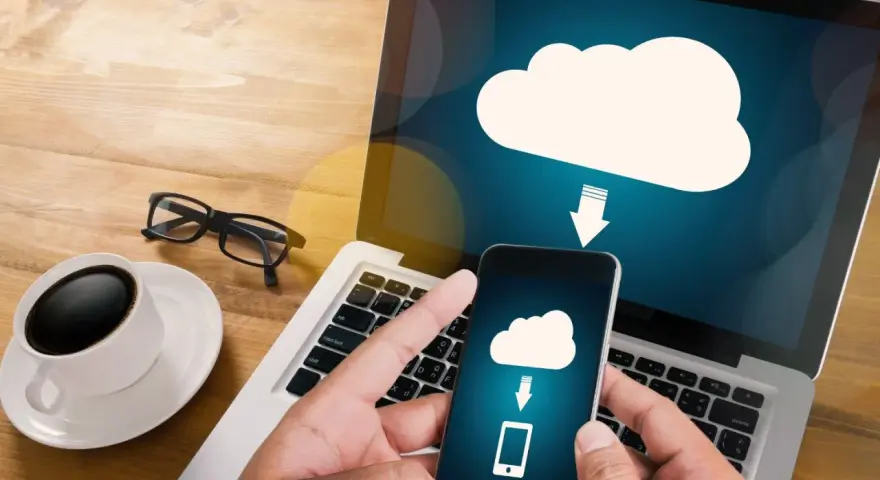 Mani che tengono uno smartphone con un'icona cloud e freccia, mostrando come salvare dati sul computer. Tazza di caffè e occhiali sul tavolo.