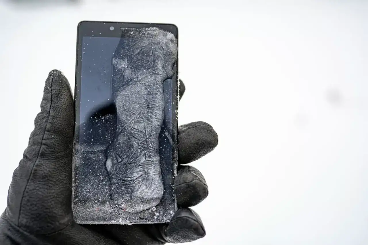 Mano con guanto nero tiene un cellulare che si impalla, coperto di ghiaccio e neve. Il freddo estremo ha bloccato il dispositivo.
